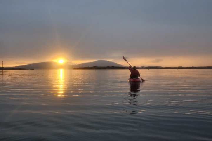 Person kayaking on calm water during sunset with mountains in the background.