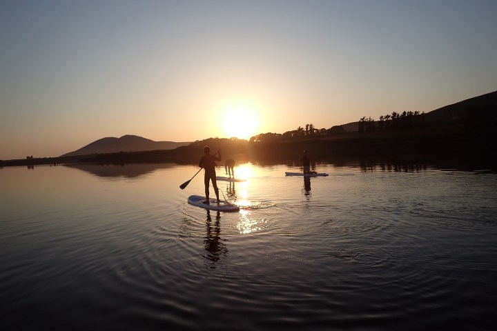 People paddleboarding on a calm lake at sunset with mountains in the background.