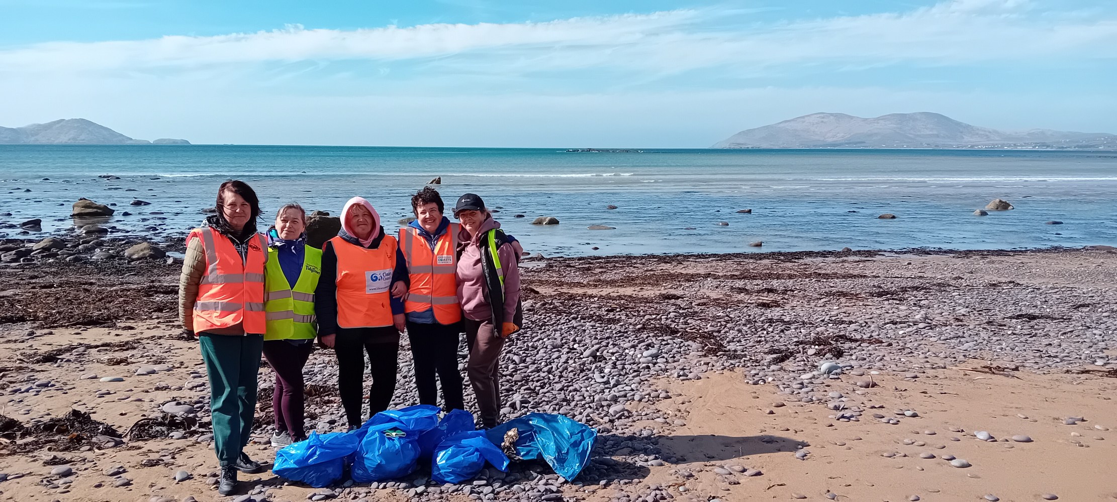 Five people in safety vests pose on a rocky beach with collected trash in blue bags.
